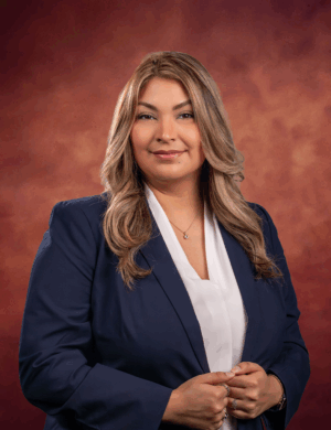 Kassandra Gonzalez, a woman with long, wavy blonde hair, poses confidently in a navy blazer and white blouse against a warm, reddish-brown studio background. She smiles gently with her hands clasped in front of her.
