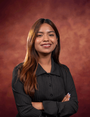 Sesilia Ponce, a woman with long brown hair wearing a black blouse, stands with her arms crossed, smiling confidently in front of a warm, reddish-brown textured background.
