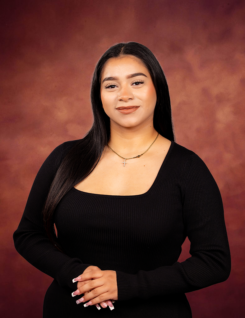 Ashley Ibarra, with long, straight black hair, wearing a black square-neck top and a gold necklace, stands against a warm brown gradient background, smiling softly with her hands clasped in front of her.