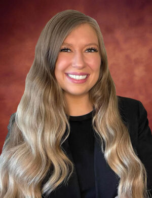 Brittany Kuusisto, with long, wavy blonde hair, wearing a black top and blazer, smiles in front of a warm, reddish-brown gradient background.