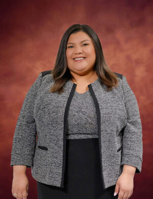 Marisa Ayala, a woman with long dark hair, wears a gray blazer over a patterned top and black skirt, standing and smiling against a reddish-brown studio background.