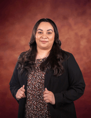 A woman with long dark hair wearing a black blazer over a floral top stands in front of a warm, reddish-brown background, facing the camera and smiling gently.