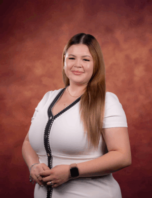 Brianna Valadez, a woman with long, light brown hair, smiles in a white dress with black trim before a reddish-brown gradient backdrop. She wears a smartwatch and bracelets on her left wrist.
