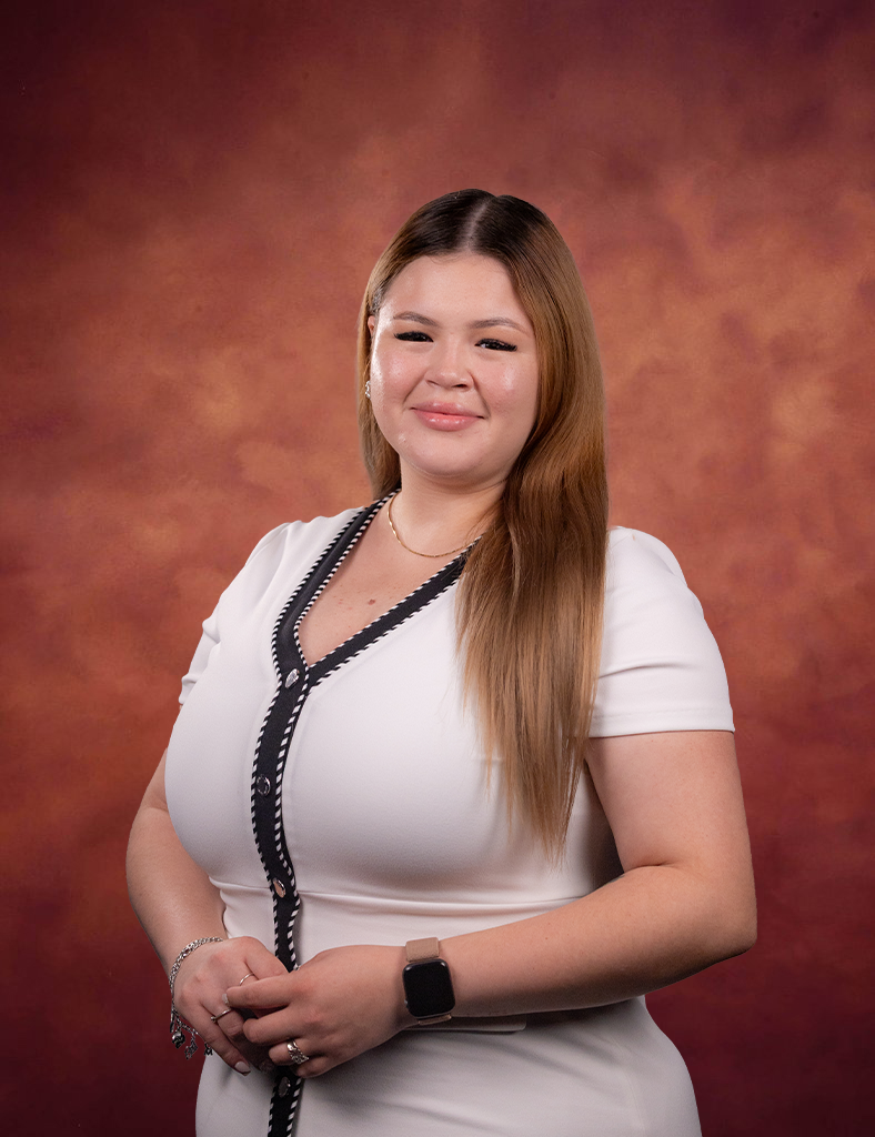 Brianna Valadez, a woman with long, light brown hair, smiles in a white dress with black trim before a reddish-brown gradient backdrop. She wears a smartwatch and bracelets on her left wrist.