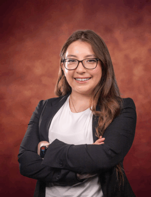 Jaqueline Gonzalez, a woman with long brown hair and glasses, wearing a black blazer and white top, stands smiling with arms crossed in front of a warm, reddish-brown background.