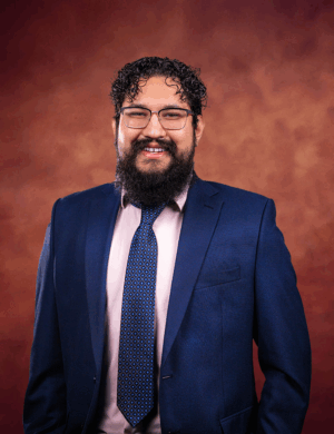 Jerry Guerra, a man with curly hair, a beard, and glasses, is smiling in a navy blue suit, light pink shirt, and patterned blue tie as he stands before a warm brown gradient background.