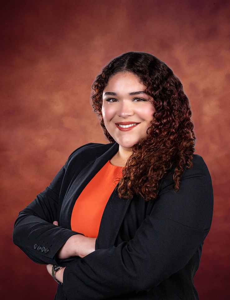 A woman with curly brown hair, wearing a black blazer over an orange top, smiles confidently with her arms crossed against a warm, reddish-brown studio background.