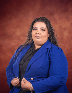 A woman with long, wavy hair wears a royal blue blazer over a black top, standing in front of a warm, brown gradient background. She faces the camera with a calm expression, holding her blazer with both hands.