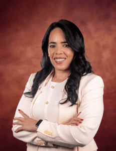 Jessica Rodriguez, a woman with long black hair wearing a cream blazer and white top, stands with arms crossed, smiling confidently against a warm brownish-red background.