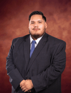Jose Luis Meza, in a dark suit, light blue shirt, and blue patterned tie, stands before a warm brown gradient background, looking at the camera with a neutral expression and his hands clasped in front.