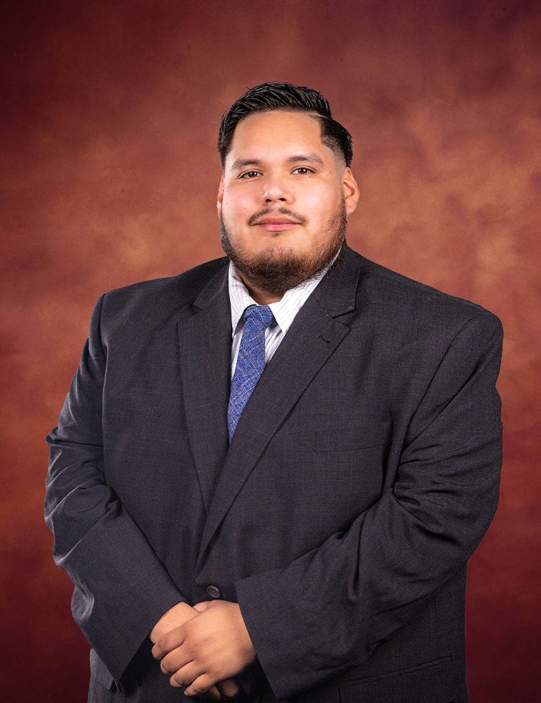 Jose Luis Meza, in a dark suit, light blue shirt, and blue patterned tie, stands before a warm brown gradient background, looking at the camera with a neutral expression and his hands clasped in front.