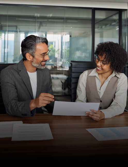Two people in business attire sit at a desk, smiling and discussing a document. Papers are spread out on the wooden table, as Personal Injury & Criminal Defense Attorneys meet in a glass-walled office in the background.
