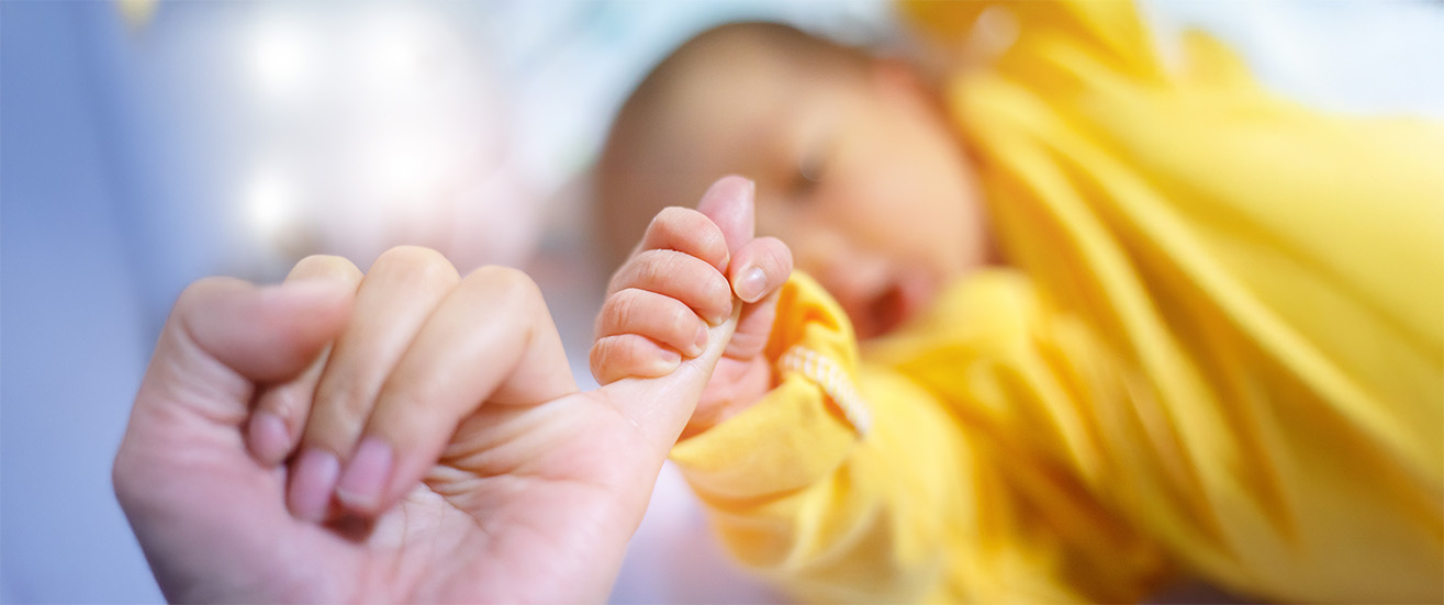 A baby in a yellow outfit, born through a Gestational Agreement, grasps an adult's finger with its tiny hand, lying on a soft surface with a blurred background.