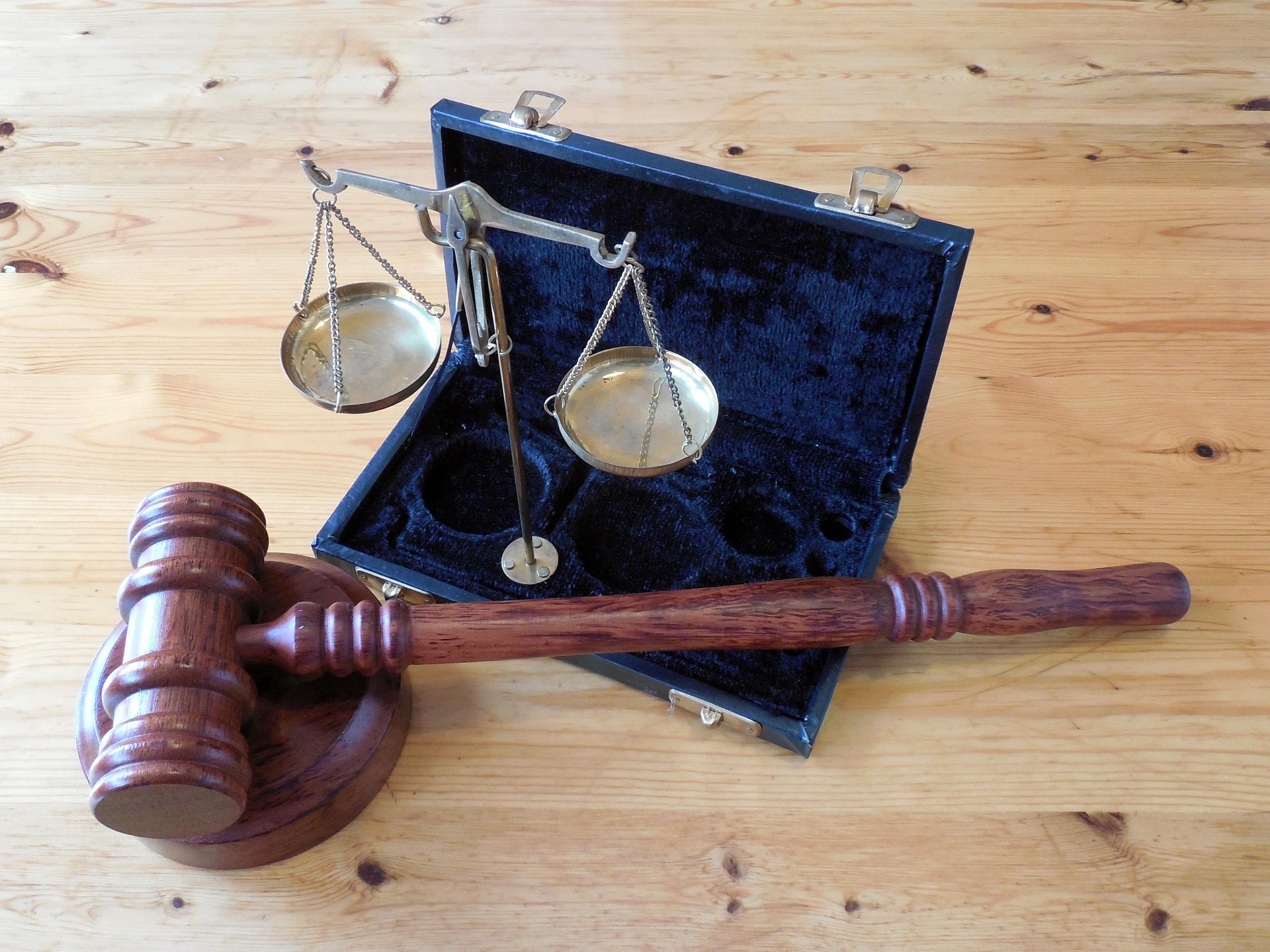 A wooden judge’s gavel and sound block rest on a table beside brass balance scales in an open black case lined with velvet, evoking the seriousness of a domestic violence charge.