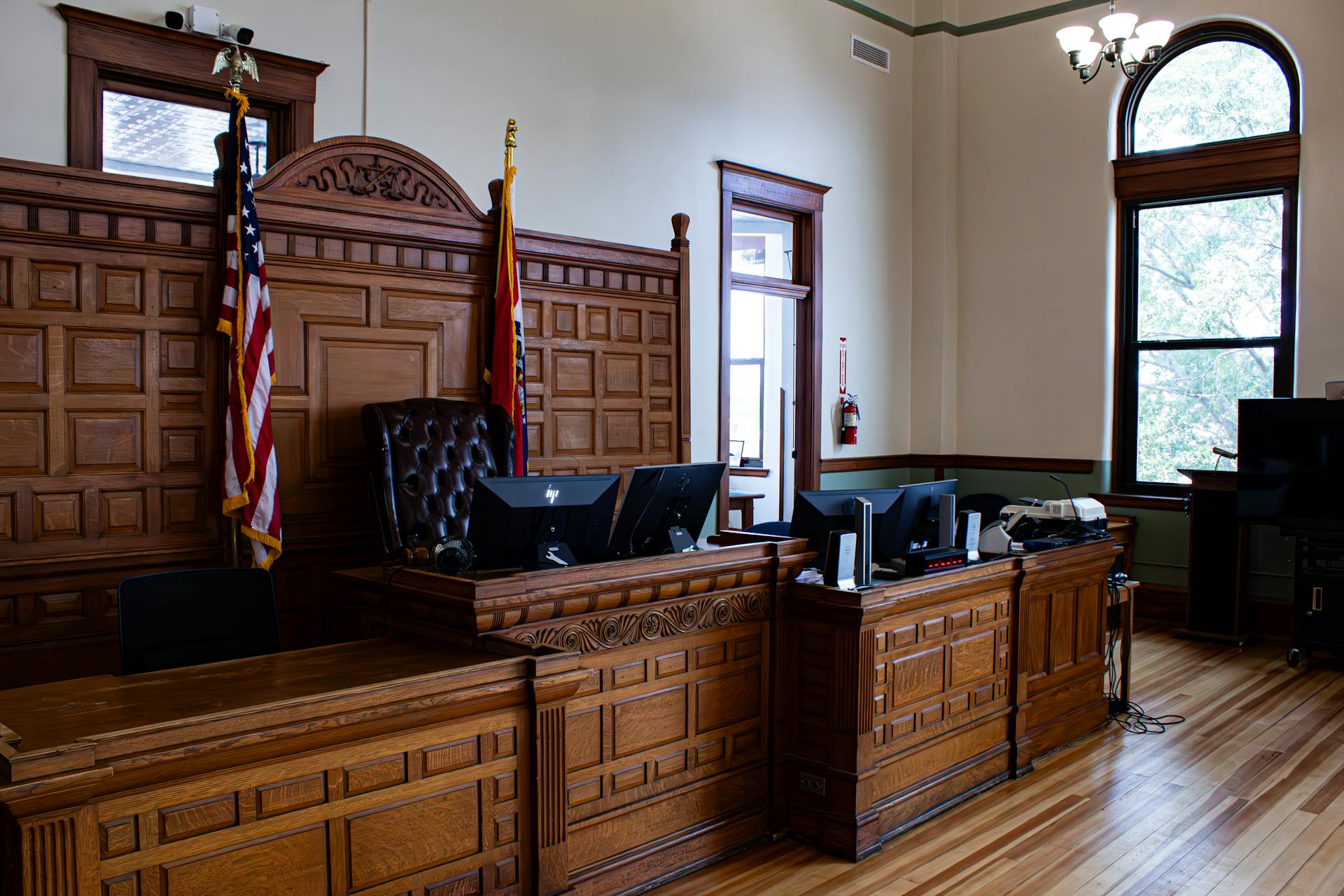 A courtroom with wooden paneling, a judge’s bench for bench trials, American and state flags, computer monitors, and large windows letting in natural light.