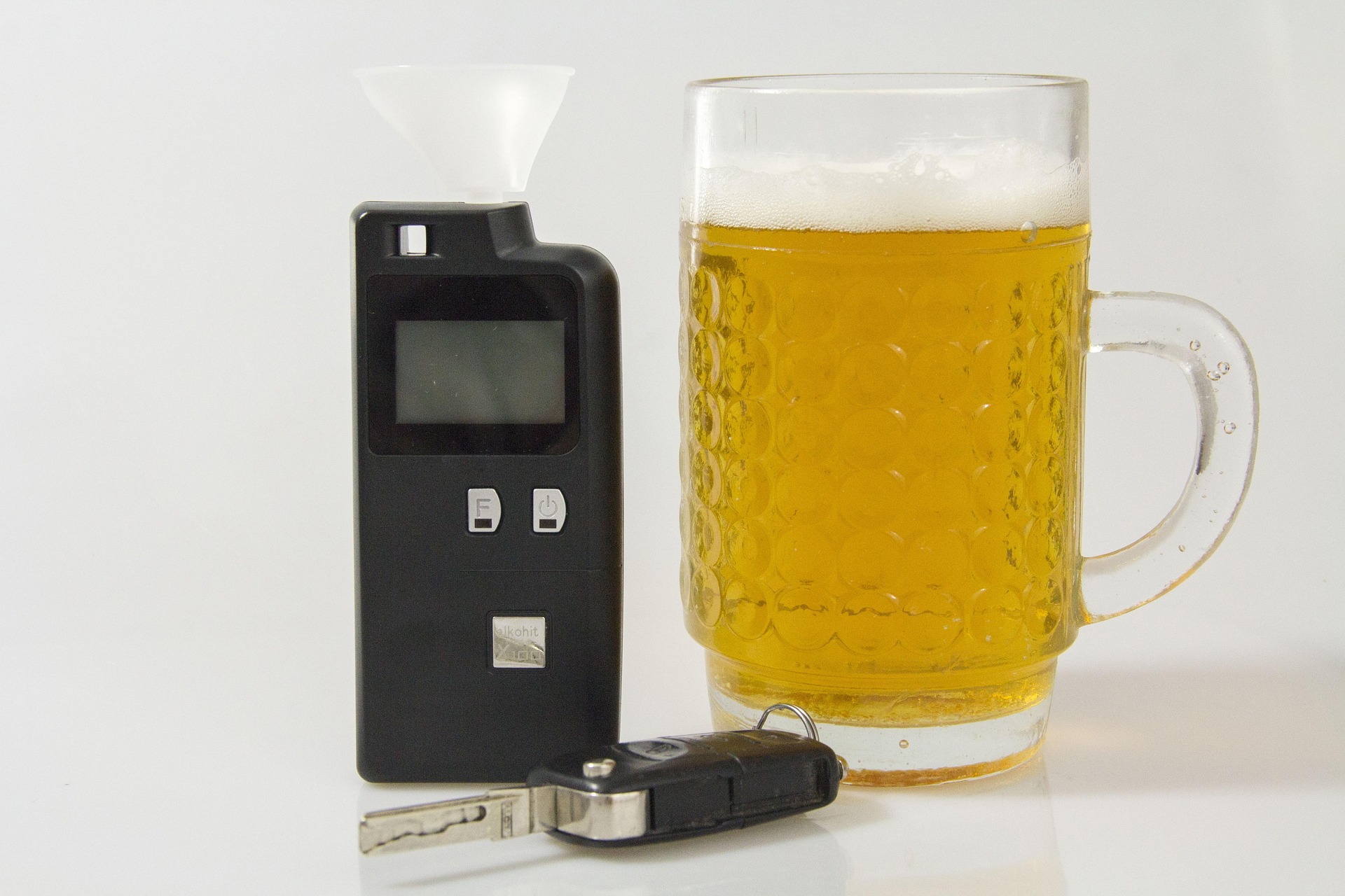 A breathalyzer, a mug of beer, and a car key rest on a white surface