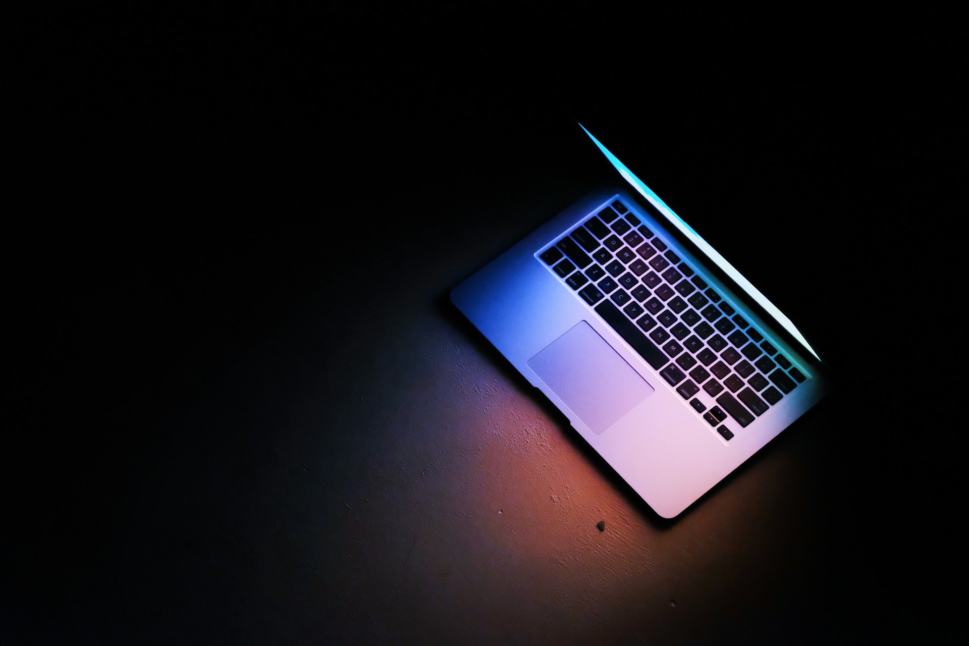 A partially open laptop sits on a dark surface, illuminated by blue and purple light from its screen, creating a dramatic and minimalistic scene that subtly hints at the hidden dangers of revenge pornography.