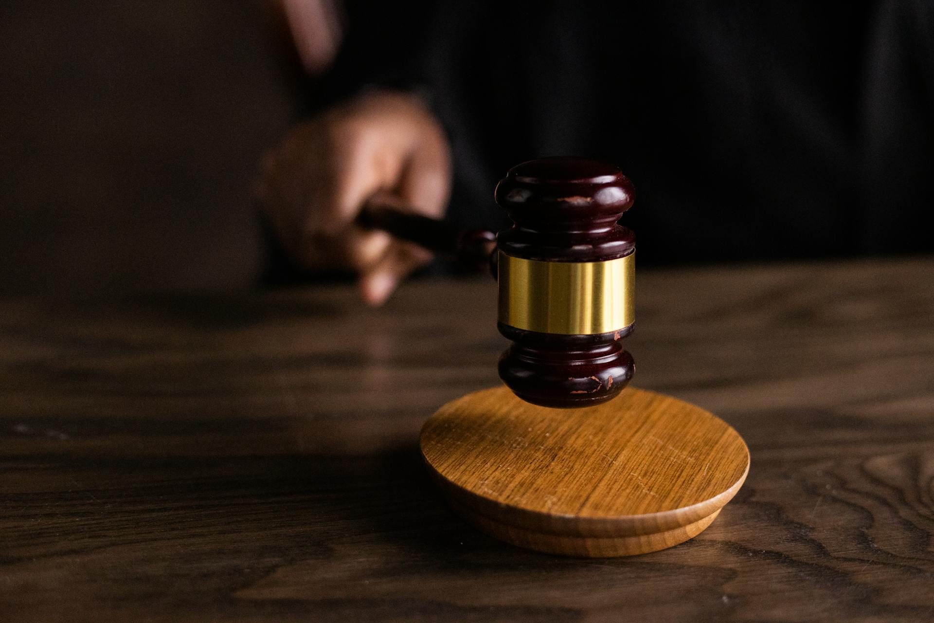A hand holding a wooden judge’s gavel poised above a round wooden sound block on a dark wooden table, symbolizing law, justice, and the weight of decisions involving criminal negligence.