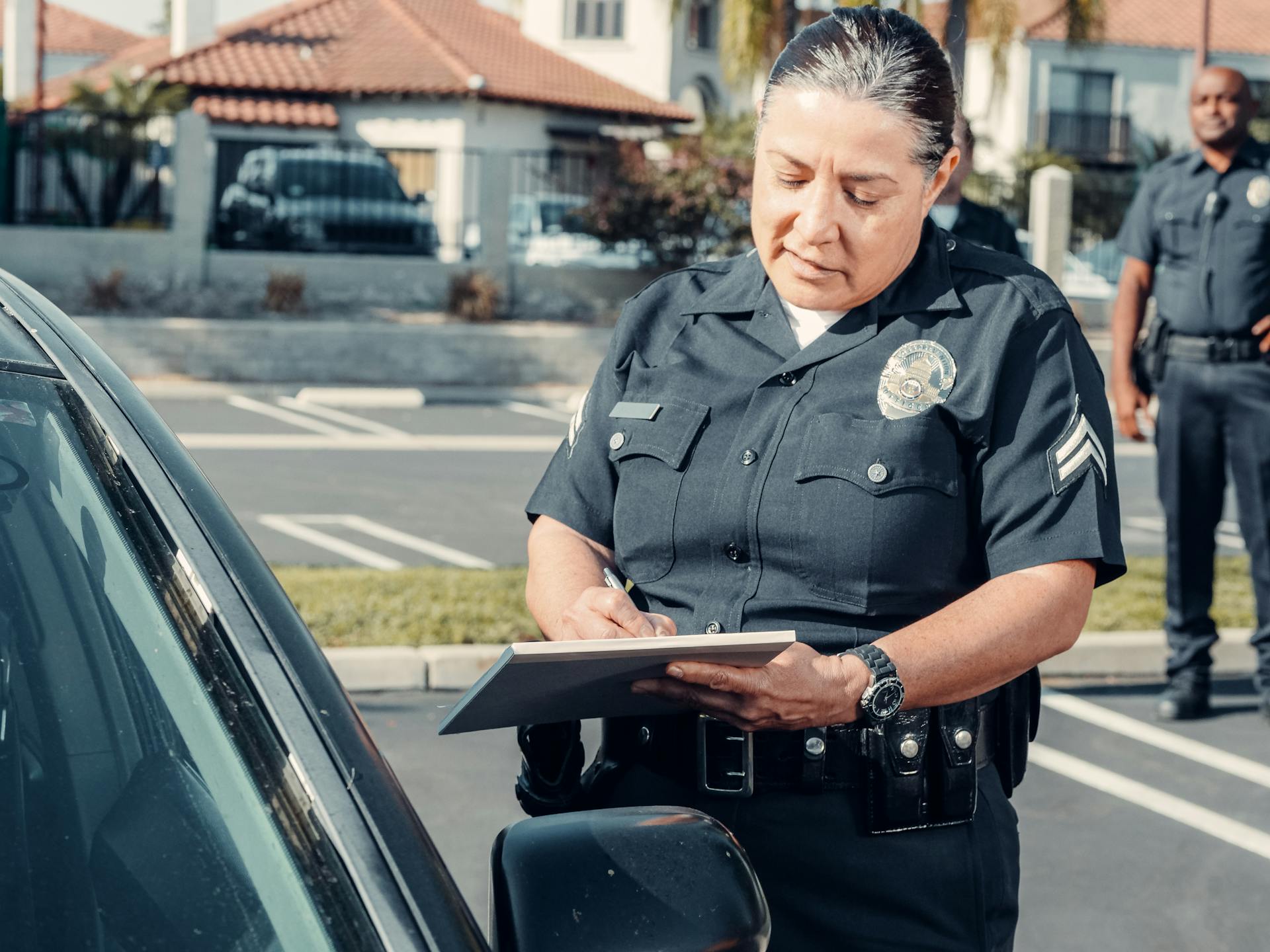 During a traffic stop, a police officer stands beside a parked car, writing on a notepad, while another officer observes in the background in a parking lot with houses visible behind them.