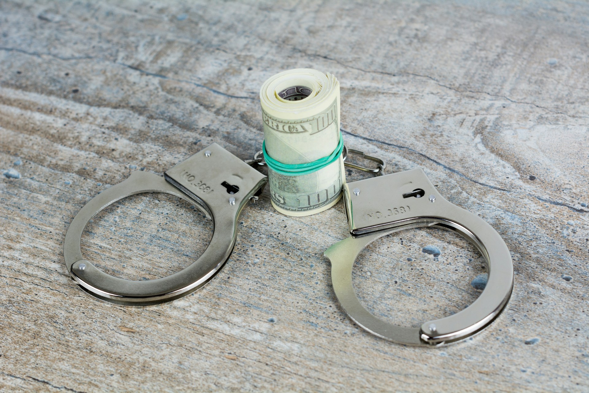 A roll of U.S. dollar bills secured with a rubber band is placed between open metal handcuffs on a wooden surface, hinting at bankruptcy fraud charges in Texas.