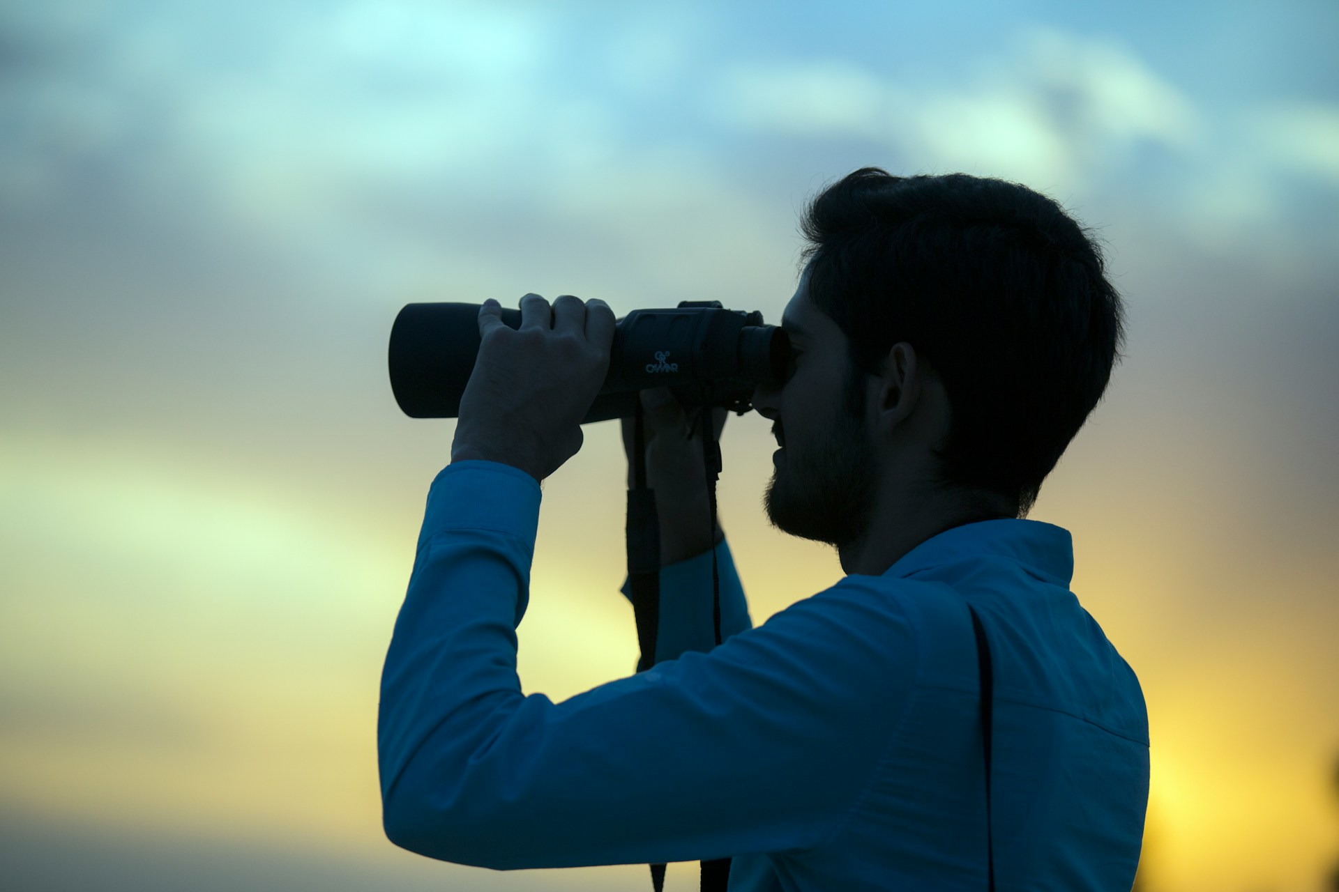 A person in a blue shirt looks through binoculars at sunset, with a colorful sky in the background—perhaps contemplating how to fight a stalking charge in Texas with the right defense.