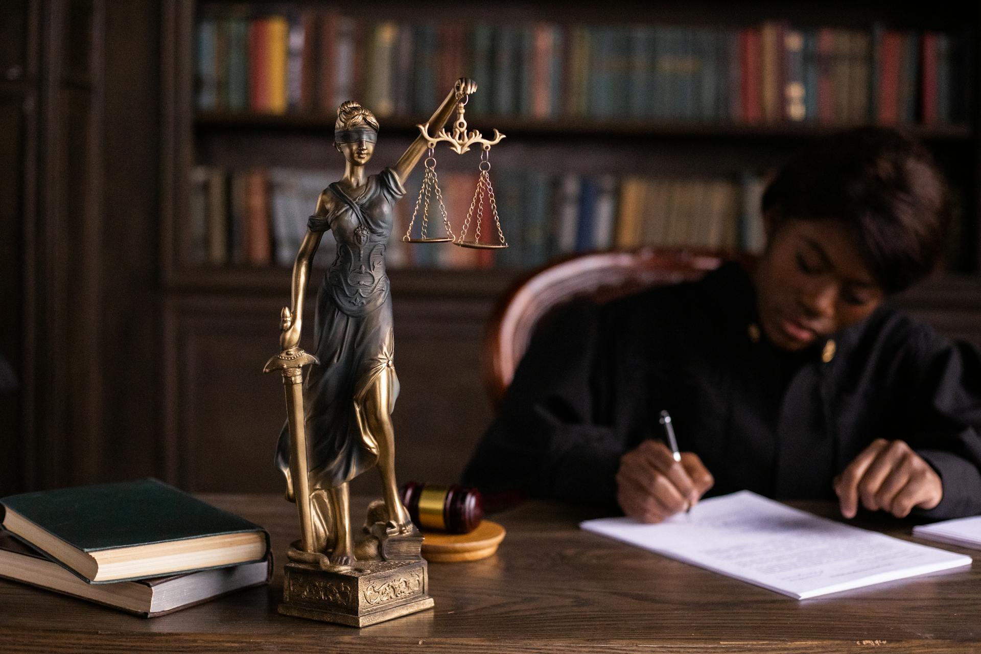 A statue of Lady Justice stands on a desk beside books, a gavel, and a restraining order, while a person in a judge’s robe writes on a document in the background, with shelves of books behind them.