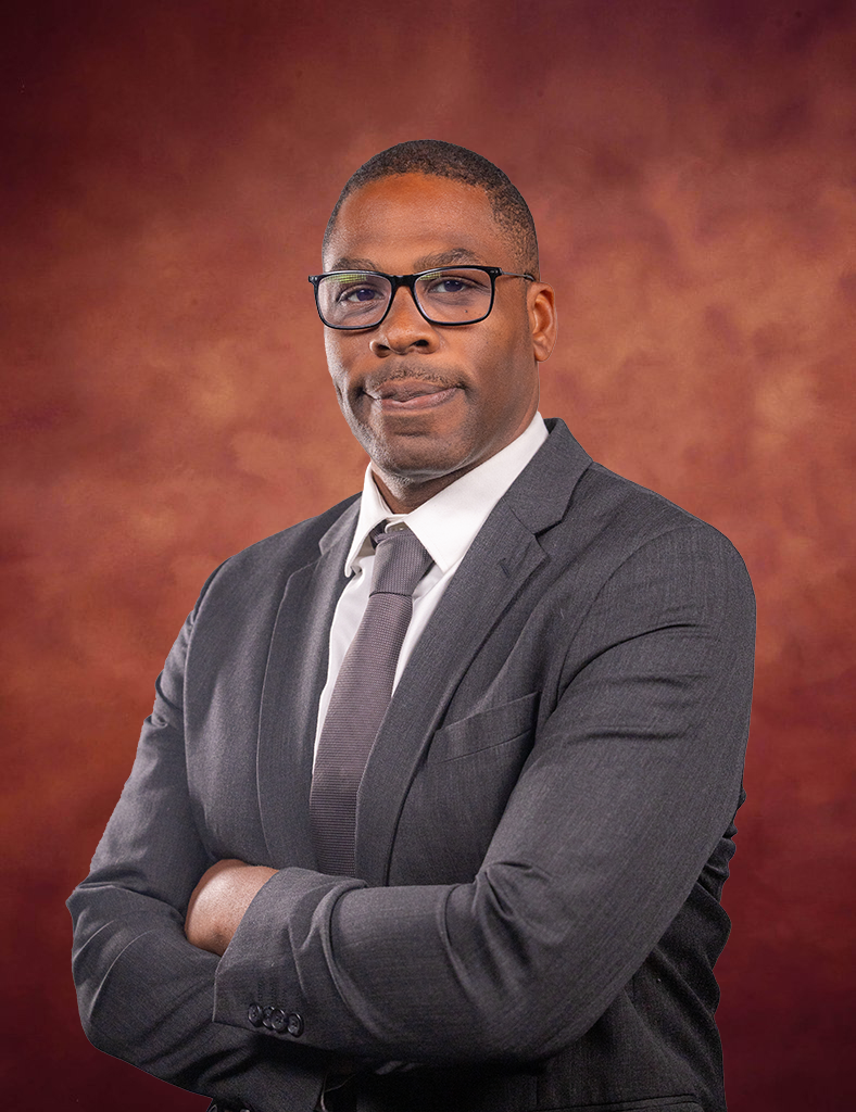 A man wearing glasses, a dark suit, white shirt, and dark tie poses with arms crossed in front of a reddish-brown, softly textured background. He looks confidently at the camera.