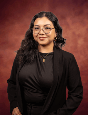 A woman with long wavy dark hair, wearing glasses, gold hoop earrings, a gold necklace, and a black blazer over a black top, stands against a reddish-brown gradient background, smiling slightly.