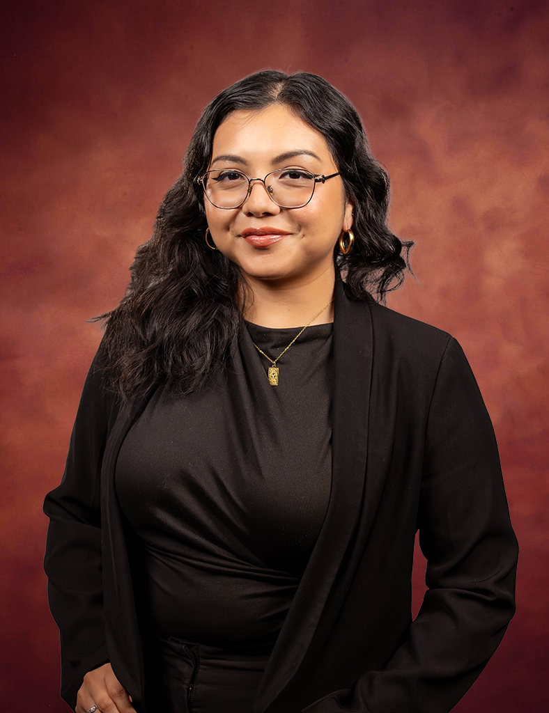 A woman with long wavy dark hair, wearing glasses, gold hoop earrings, a gold necklace, and a black blazer over a black top, stands against a reddish-brown gradient background, smiling slightly.