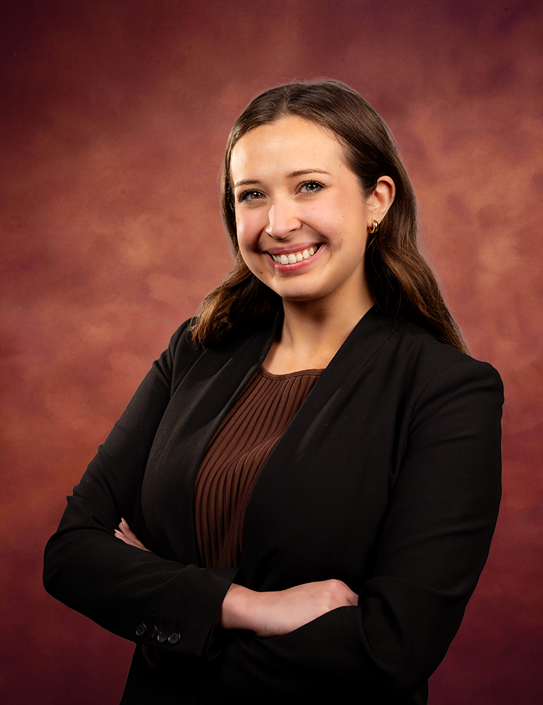 Gabriella Borrego, a woman with long brown hair, wearing a black blazer and brown top, smiles confidently with arms crossed in front of a warm, reddish-brown studio background.