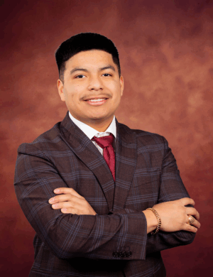 Jakob Chamberlain, a young man in a plaid suit and red tie, stands with arms crossed, smiling confidently against a warm brown studio backdrop.