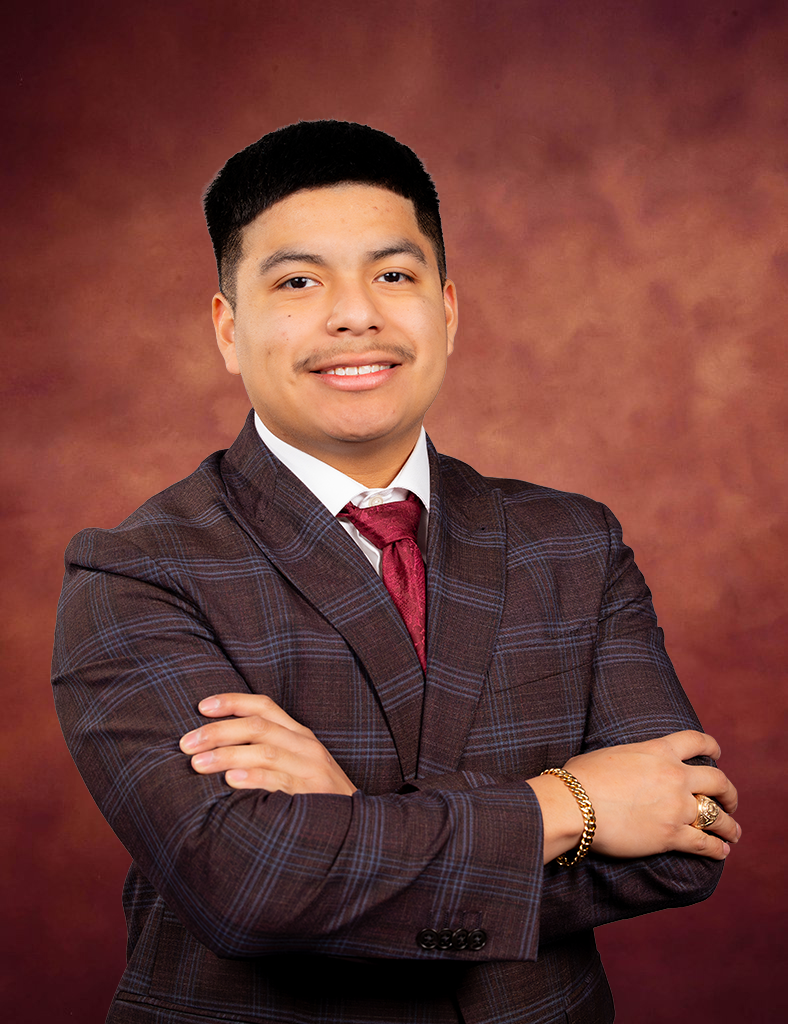 Jakob Chamberlain, a young man in a plaid suit and red tie, stands with arms crossed, smiling confidently against a warm brown studio backdrop.