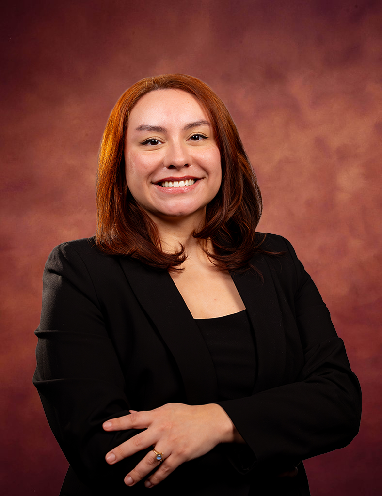 A woman with straight, shoulder-length auburn hair smiles confidently with arms crossed, wearing a black blazer and top, in front of a reddish-brown studio backdrop.