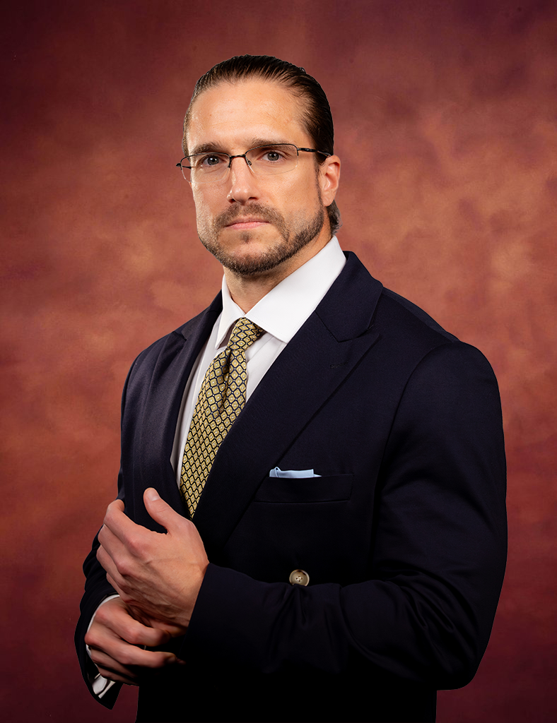 Adam Stewart, in a dark suit, patterned tie, and glasses, stands against a mottled brown backdrop. He looks directly at the camera with a serious expression, one hand adjusting his jacket.
