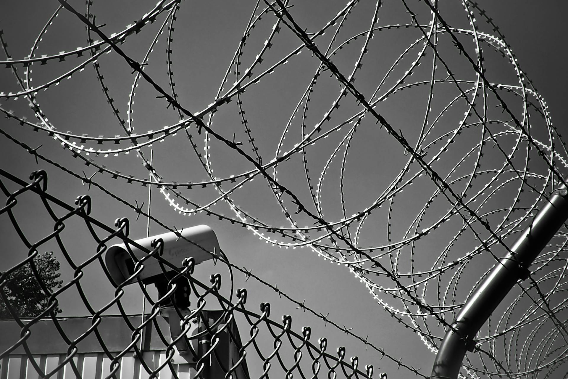 Barbed wire and chain-link fence with a security camera behind it, all under a clear sky. The image evokes high security, with barriers as unyielding as a statute of limitations—firmly enclosing what lies within.
