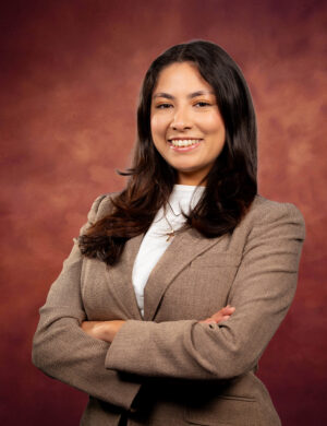 A woman with long dark hair, wearing a brown blazer and white top, stands confidently with arms crossed, smiling in front of a reddish-brown, textured background.