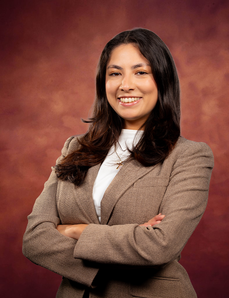 A woman with long dark hair, wearing a brown blazer and white top, stands confidently with arms crossed, smiling in front of a reddish-brown, textured background.