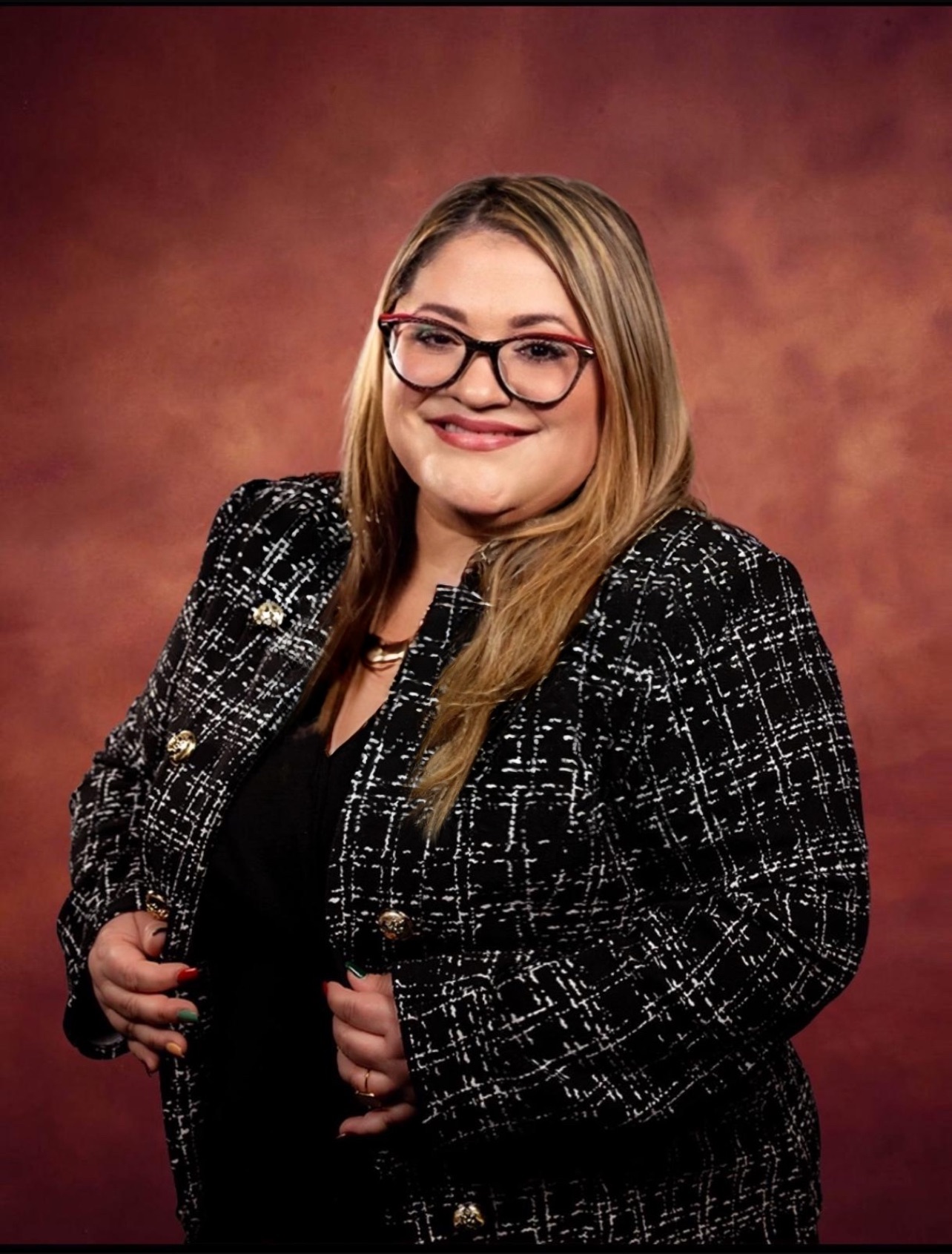 A woman with long blonde hair and glasses smiles at the camera. She is wearing a black and white patterned blazer over a black top, standing against a warm brown background.