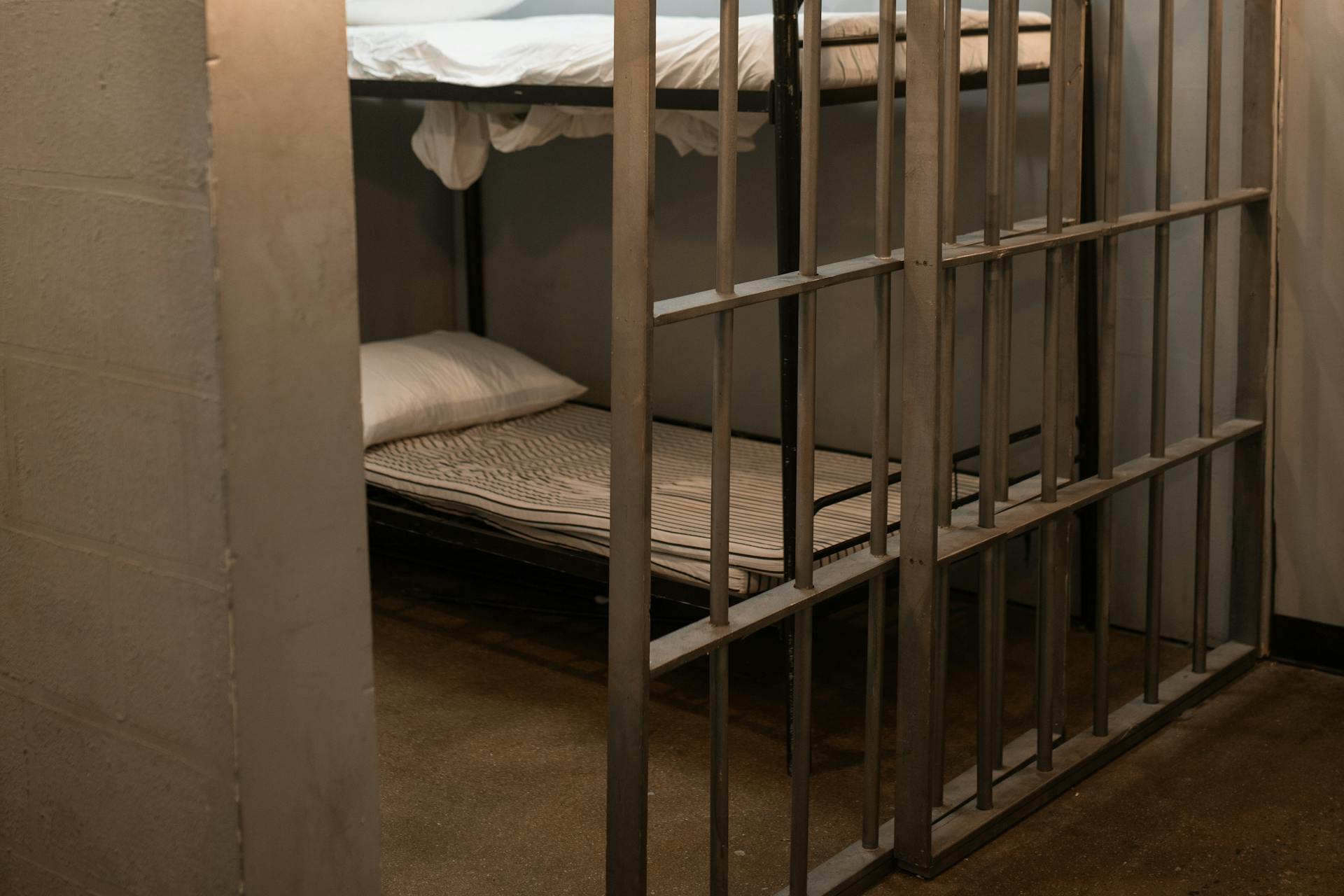 A small jail cell with metal bars, often used to detain individuals accused of felony theft, contains two bunk beds with white sheets and pillows, and a plain concrete floor.