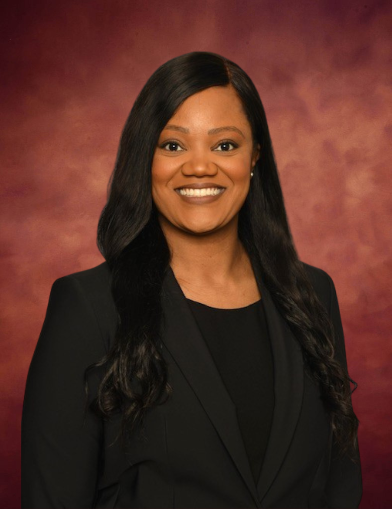 Erika T. O’Neal, a woman with long, dark hair wearing a black blazer and top, smiles warmly in front of a reddish-brown background.