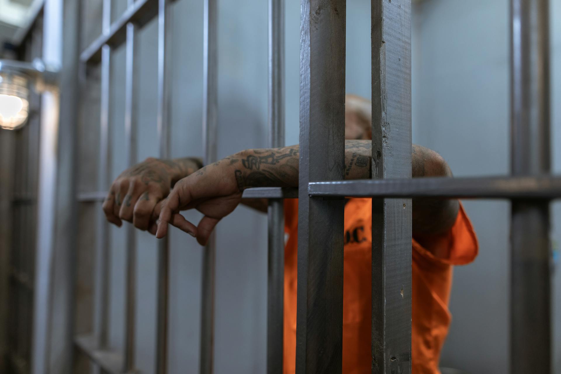 A person in an orange prison uniform, jailed for domestic violence, stands behind bars with tattooed arms resting through them. Their face is obscured and the setting appears to be a dimly lit jail cell.
