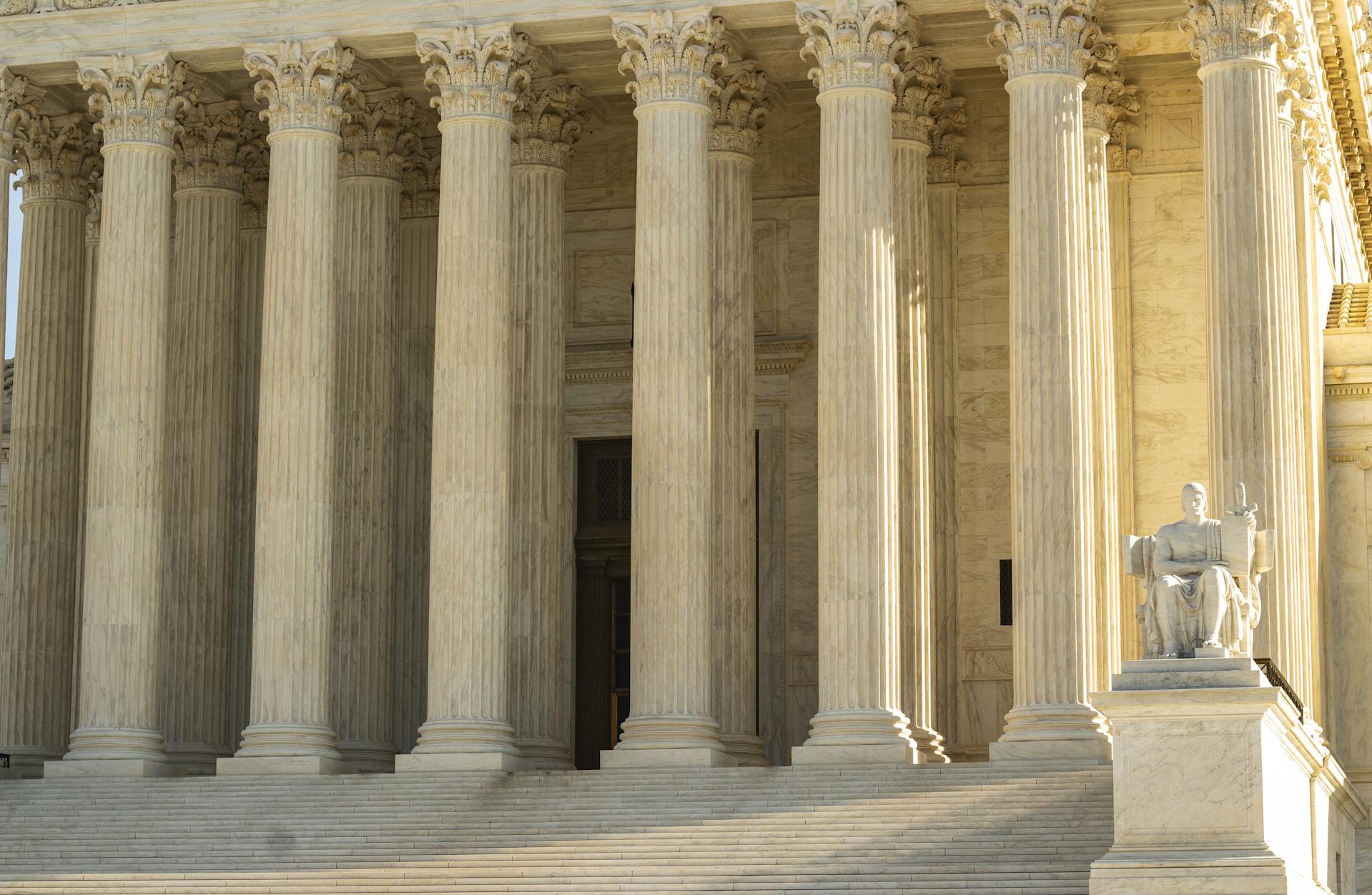 Wide stone steps lead up to the entrance of a grand Texas building with tall marble columns and a seated statue holding a book and staff on the right. Sunlight casts shadows across the impressive structure.