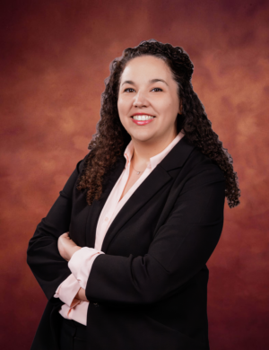 Maria Vargas, a woman with curly hair wearing a black suit and light pink blouse, stands smiling with arms crossed in front of a brown gradient background.