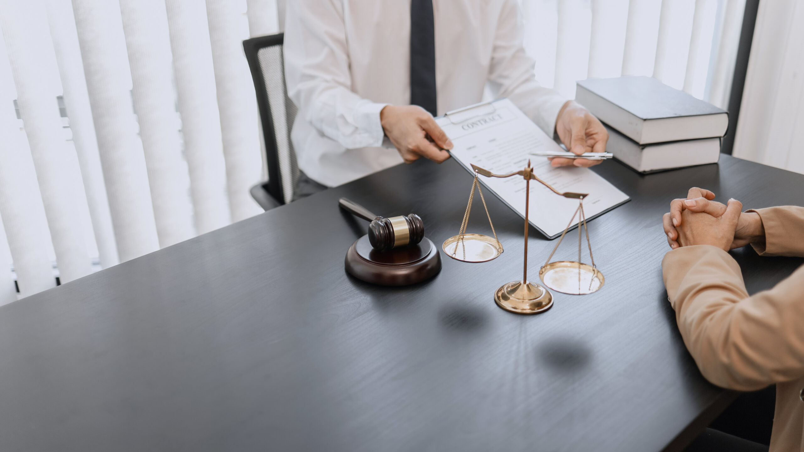A Texas attorney sits at a desk holding a contract and pointing with a pen while talking to a client. On the desk are a gavel, scales of justice, and stacked books. The client’s hands are visible across the desk, seeking an innocent verdict.