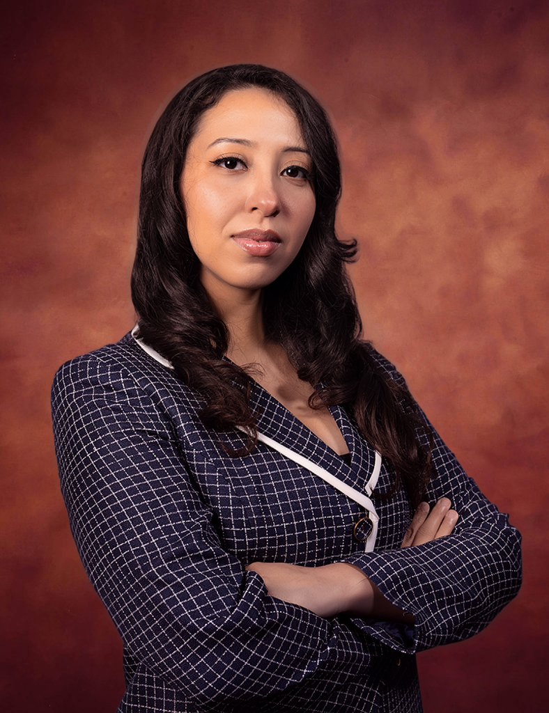 A woman with long dark hair wearing a blue and white checkered blazer stands with arms crossed against a reddish-brown studio background, looking confidently at the camera—an image reminiscent of an Eliud Romano portrait.