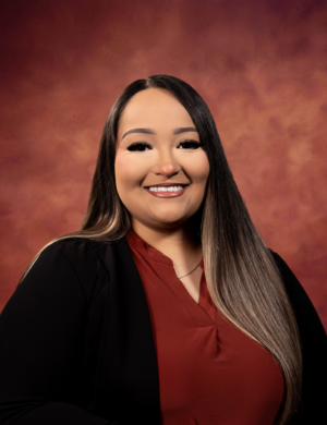 Julia Martinez, with long, straight hair and a warm smile, poses against a reddish-brown studio backdrop. She is wearing a maroon top and a black blazer.