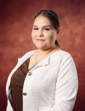 Bianka Garza, a woman with light skin and straight, pulled-back hair, poses in front of a warm brown backdrop. She wears a white textured blazer over a brown top and smiles softly at the camera.