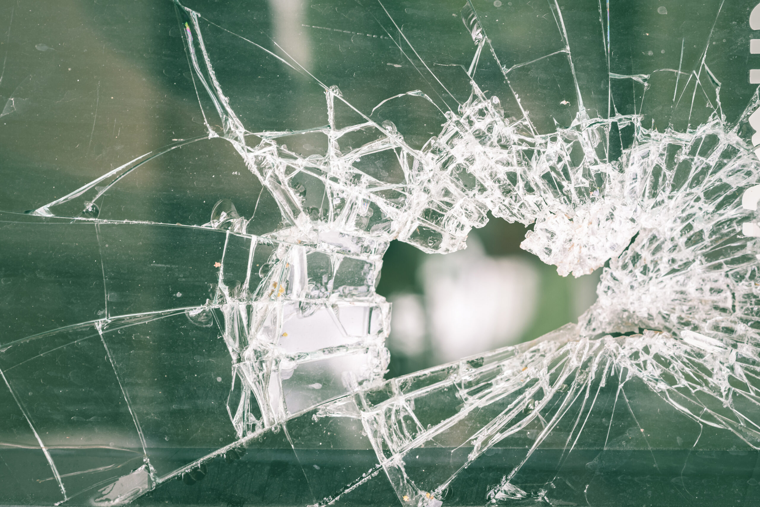 A close-up of a shattered glass pane with a hole in the center and cracks radiating outward, set against a blurred green background—imagery often linked to a criminal mischief charge.